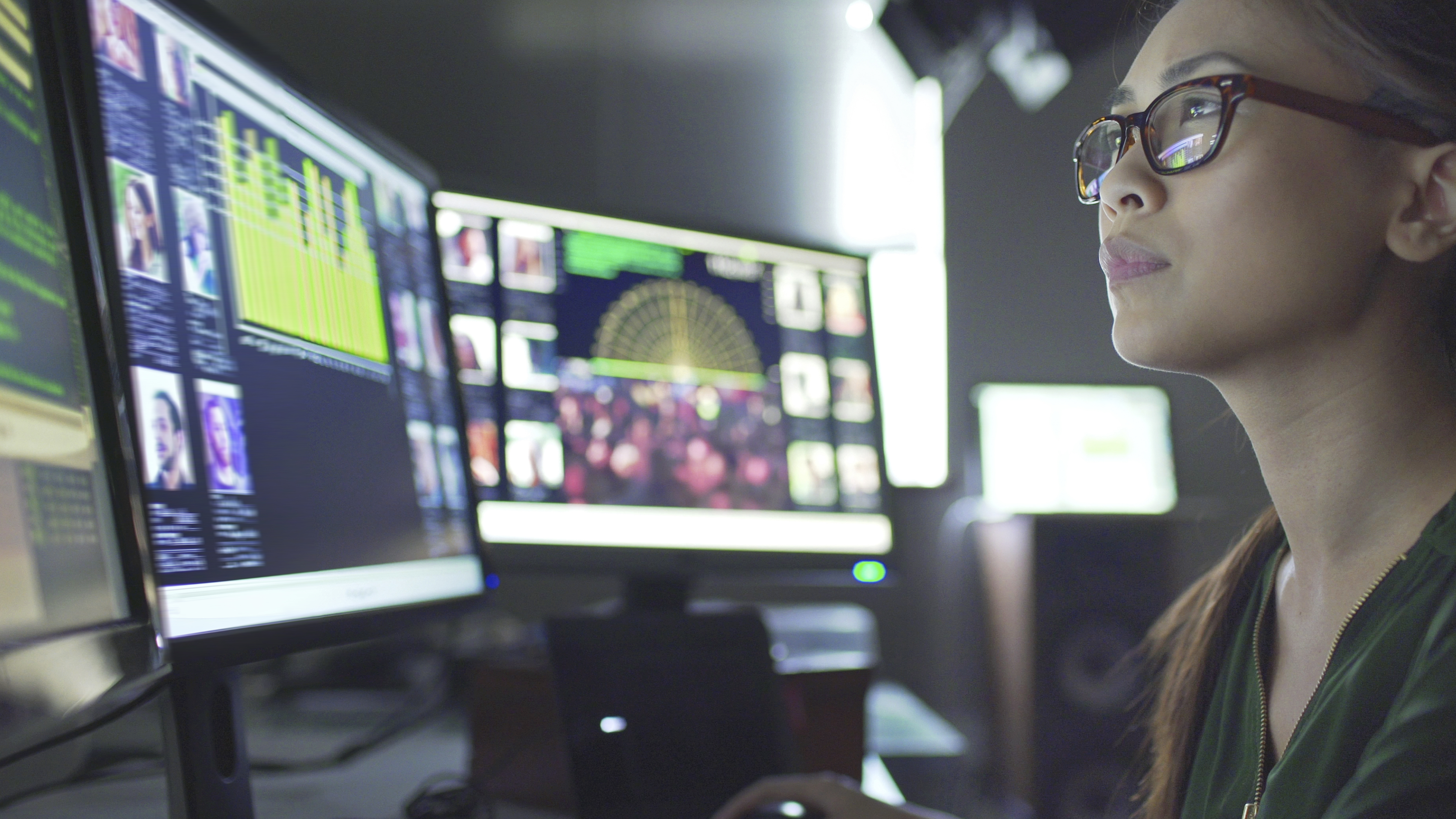 young asian woman sitting down at her desk where she’s surrounded by 3 large computer monitors displaying out of focus images of people as thumbnails; crowds; graphs and scrolling text.